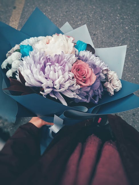 A floral bouquet featuring large, soft purple chrysanthemums, a single pink roses, and accent flowers in shades of white and blue. The flowers are arranged tightly in a bouquet with fresh, vibrant pet