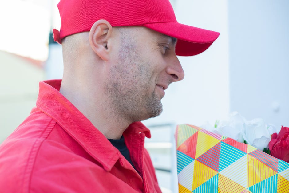 A delivery worker wearing a red cap and matching red jacket is seen holding a floral gift wrapped in colorful, geometric-patterned paper with a mix of yellow, blue, red, and white shades. The bouquet 