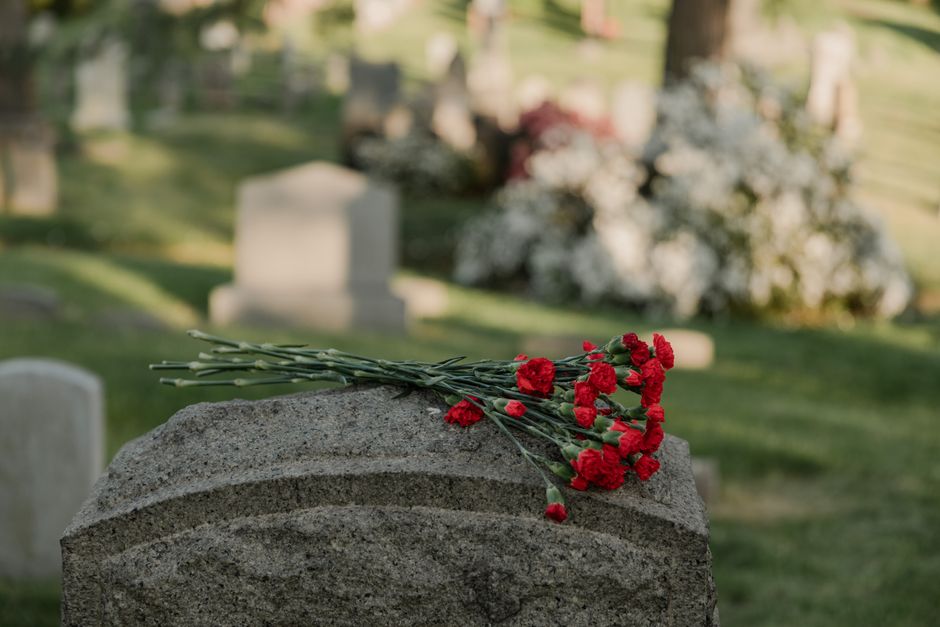 A bouquet of fresh red carnations with green stems and leaves, resting on a grey granite grave monument in a cemetery. The flowers are arranged casually, with some stems slightly overlapping each othe