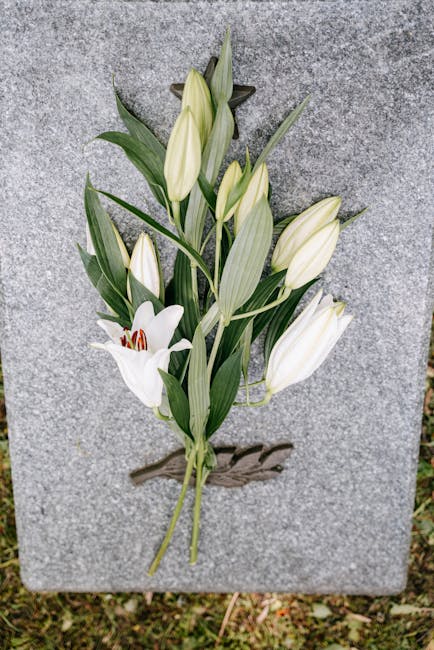 A single bouquet of white lilies and unopened lily buds with long green leaves, arranged on a polished gray granite memorial stone. The flowers are fresh, with delicate petals and sturdy stems, some r