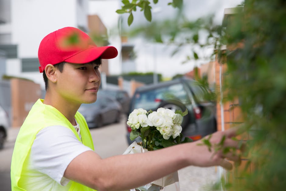 A young male florist with light skin, wearing a bright red cap and a high-visibility yellow vest over a white t-shirt, arranging a bouquet of fresh white hydrangeas with dark green leaves. He is stand