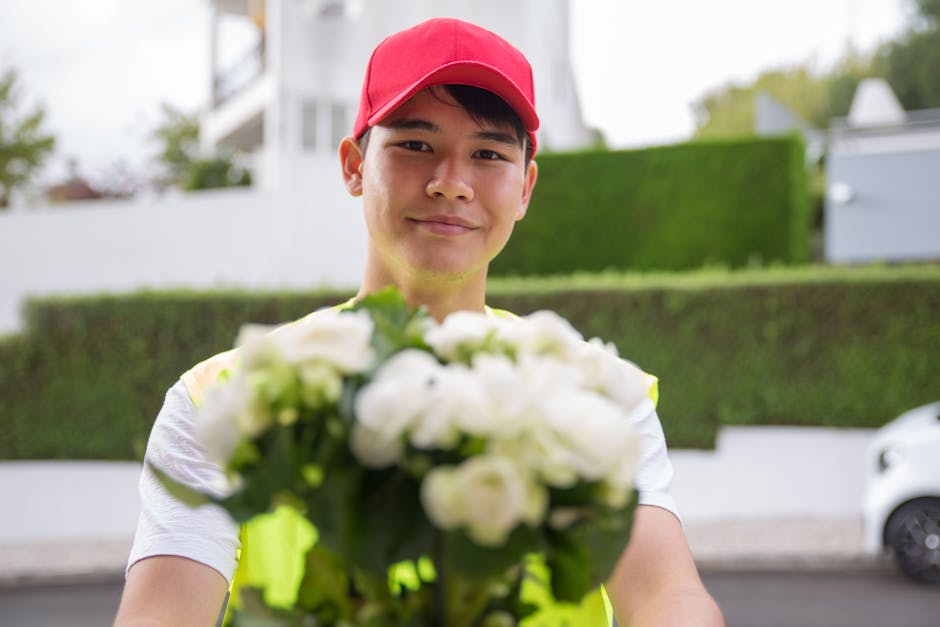 A delivery person wearing a red cap and a reflective vest extends a bouquet of fresh white roses and green foliage towards the camera, smiling gently. The bouquet is held with both hands and is wrappe